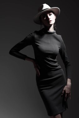 Zarif bir moda. Dar gri elbiseli, siyah şapkalı, gri stüdyo arka planında poz veren zarif, ince bir bayan..