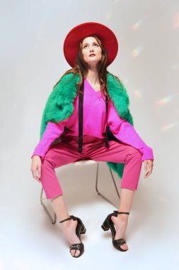 Parlak fuşya kıyafetleri, yeşil kürk manto ve sandalyenin üzerinde zarif bir şapka ile poz veren abartılı, güzel bir kadının tam boy portresi. Modada parlak renkler. Hafif bir arkaplanda stüdyo çekimi.