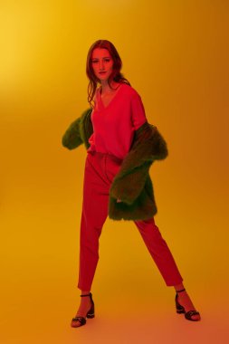 Yüksek moda çekimi. Güzel bir mankenin tam boy portresi. Parlak giysiler içinde sarı arka planda kırmızı ışıkta poz veriyor..