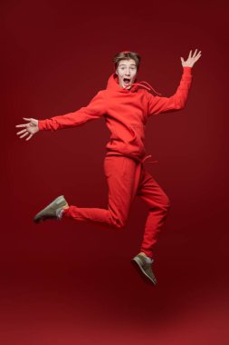 Mutluluk, neşeli duygular, başarı. Kırmızı eşofmanlı heyecanlı genç çocuk sevinçten zıplayıp çığlık atıyor. Kırmızı arka planda tam boy stüdyo resmi.