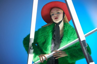 Yüksek moda ve tarz. Parlak makyajlı, sarı saçlı, şık yeşil kürklü ve neon lambaların arasında kırmızı şapkalı çarpıcı bir manken kız. Mavi arkaplanda stüdyo resmi. 