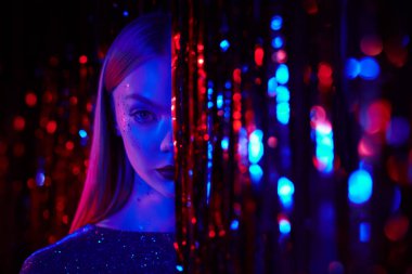 Gece kulübü, disko, neon ışıklar. Parlak makyajlı ve gece elbisesi parıldayan şık sarışın bir kız kırmızı folyo perdelerin arasında duruyor. Göz kamaştırıcı bir parlaklık, moda ve tarz. Boşluğu kopyala. 