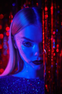 Parlak makyajlı ve gece elbisesi parıldayan şık sarışın bir kız kırmızı folyo perdelerin arasında duruyor. Gece kulübü, disko, neon ışıklar. Parıltı, moda ve tarz. 
