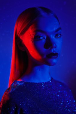 Gece kulübü, disko, neon ışıklar. Parlak makyajlı şık bir sarışın kız ve parlak bir gece elbisesi mavi arka planda duruyor. Göz kamaştırıcı ışık, moda ve tarz. 
