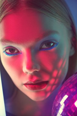 Parlak makyajlı güzel bir sarışın kız, neon lambaların ışığında parlayan bir disko topuyla poz veriyor. Stüdyo çekimi. Güzellik ve makyaj. Göz kamaştırıcı bir stil, sanatsal makyaj. Disko, gece kulübü..