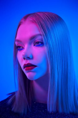 Parlak parlak makyajlı ve parlak bir gece elbisesiyle göz kamaştırıcı sarışın kız karışık renkli ışıkta mavi bir arka planda poz veriyor. Parıltı, moda ve tarz. Saç stili.