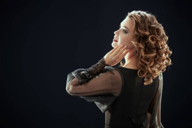 Akşam saç stili, makyaj ve moda. Siyah bir gece elbisesi, kıvırcık saç stili ve parlak akşam makyajı ile siyah bir stüdyo arkasında güzel bir genç kadın. Boşluğu kopyala.