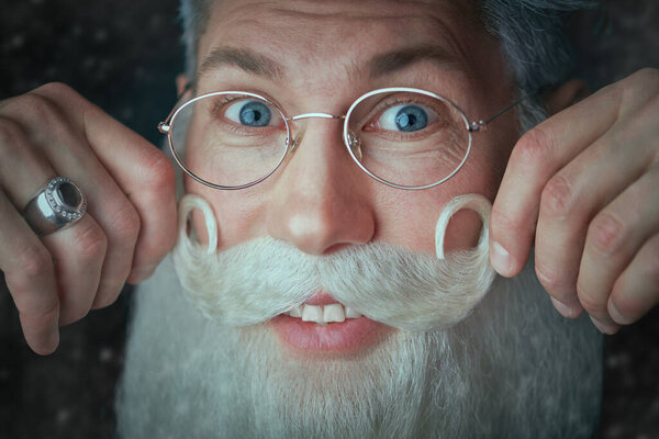 Close up shot of a well-groomed Santa Claus in glasses with elegant white beard and mustache. Dark background with snowflakes. Men's beauty, barbershop. Christmas and New Year.