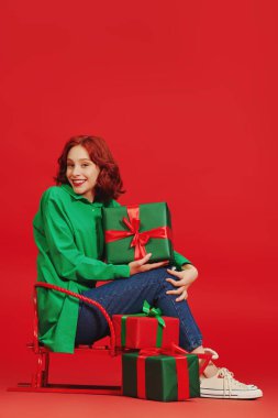 Sevimli, kızıl saçlı, hediye kutularıyla kızakta oturan bir kız. Fotokopi alanı olan kırmızı bir arkaplan. Noel satışları. Hediye zamanı. Noel ve Yeni Yıl Hediyeleri. 