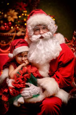Parlak bir şenlik elbisesi ve Noel Baba şapkası giymiş küçük bir kız Noel Baba 'nın kucağında oturuyor. Mutlu Noeller ve mutlu yıllar.!
