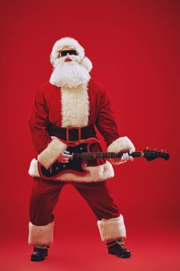 Güneş gözlüklü ve kulaklıklı modern Noel Baba elektro gitar çalıp şarkı söylüyor. Mutlu noeller. Parti zamanı. Tam yol ileri. Kırmızı arkaplan.