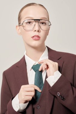 Katı makyaj ve saç stiline sahip güzel bir manken. Üç parçalı şık bir takım elbise içinde, açık arkaplanda bir stüdyoda kravat ve şık gözlükler takıyor. Moda ve iş stili. 