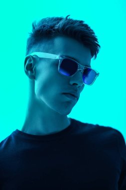Siyah uzun kollu gömlekli ve parlak yeşil-mavi arka planda poz veren havalı bir adam. Erkek modası ve tarzı. Gözlük stili.