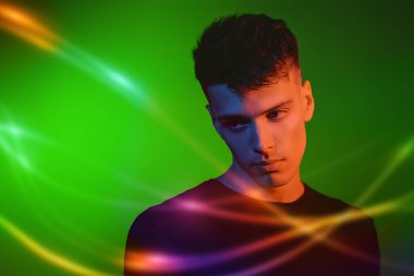 Erkek güzeli. Siyah günlük giysiler içinde yakışıklı bir esmer adamın stüdyo fotoğrafı. Farklı renklerde ışıklandırılmış yeşil bir arka plan. Gençler, duygular.