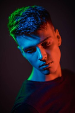 Siyah günlük giysiler içinde üzgün, yakışıklı bir esmer adamın stüdyo portresi. Koyu arkaplan ve karışık renkli ışıklandırma. Gençler. Erkek güzeli.. 