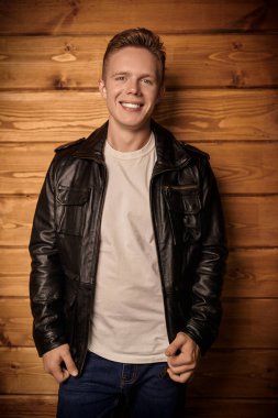 Şık deri ceketli mutlu, genç, sarışın bir adam ahşap bir duvarın yanında duruyor. Erkek modası, deri tarzı..