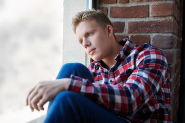 Gündelik giysiler içinde sarışın bir adam pencere pervazındaki çatı katında oturur ve pencereden dışarı bakar. İnsanlar, yaşam tarzı, duygular. Boşluğu kopyala.