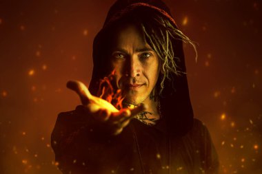 Etnik giyinmiş, kapüşonlu bir erkek şaman elini uzatıyor ve içinden çıkan ateşle sizi gizemli dünyasına davet ediyor. Kıvılcımlı koyu kırmızı arka plan. Cadılar Bayramı, Fantezi.