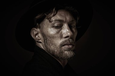 Art portrait of a mature man with beard and moustache dressed in black coat and black hat looking pensively down. Dark key portrait on a black background. Male beauty.  clipart