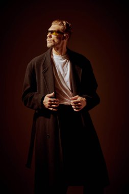 Erkek modası ve güzellik. Uzun siyah ceketli ve güneş gözlüklü zarif giyimli olgun bir adam loş ışıklandırmalı karanlık bir stüdyonun arka planında duruyor.. 