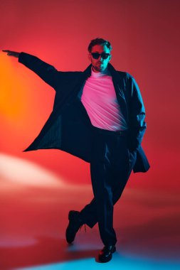 Zarif kıyafetler ve kırmızı arka planda siyah güneş gözlüğü takan yakışıklı bir adamın moda çekimi. Karışık renkli ışıklandırma. Erkek modası.