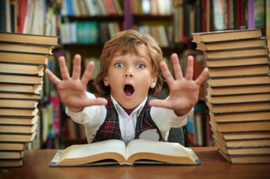 Temiz üniformalı duygusal bir öğrenci, okul kütüphanesinde kameraya korkuyla bakan kitap yığınlarının arasında oturuyor. Modern eğitim ve çocuklar.