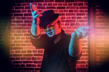 Modern kıyafetlerle, şık siyah şapkalı, sarı yuvarlak gözlüklü, kırmızı ışıkla aydınlatılmış çatı katında dans eden rapçi bir adamın portresi. Gece kulübündeki rapçi çocuk. Erkek gençlik tarzı.