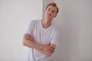 Yakışıklı genç sarışın bir adam beyaz bir duvara yaslanmış ve gülümsüyor. Mutlu insanlar, olumlu duygular. Erkek güzelliği ve sağlığı. 