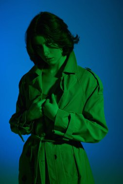Stüdyoda mavi arka planda yeşil ışık altında yağmurlukla poz veren güzel esmer kız modeli. Moda fotoğrafı.. 