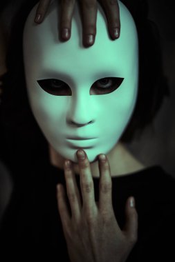 A girl in black clothes tries on a  mask-role, standing on a dark background. Human roles. Hypocrisy. Mental disorders. clipart