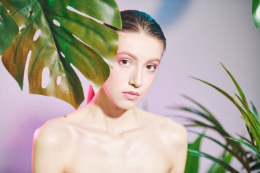 Tropik yeşilliklerin arasında duran yumuşak makyajlı güzel bir kızın portresi. Açık mor ve pembe arka plan. Orman fotoshop 'u. Spa, cilt bakımı ve kozmetik ürünleri. Güzel kız..