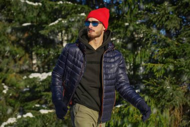 Kış kıyafetleri ve güneş gözlükleriyle yakışıklı bir adam kış parkında köknar ağaçlarının arasında yürüyor. Kış modası. 