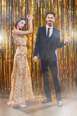 Lüks Noel partisi. Lüks gece kıyafetleri içinde yakışıklı bir adam ve güzel bir kadın parlak altın folyo perdelerin arkasında mutlu bir şekilde dans ediyorlar. Tatil modası.