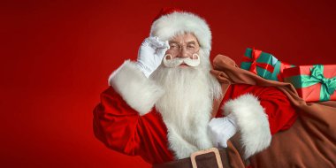Büyük bir çanta dolusu hediyeyle birlikte nazik bir Noel Baba portresi. Fotokopi alanı olan şenlikli kırmızı stüdyo. Noel ve Yeni Yıl.