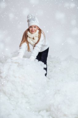 Çocuklar için kış modası. Ilık örgü elbiseli güzel neşeli kız beyaz karlı arka planda büyük bir kartopu yapıyor. Kış aktiviteleri ve tatiller. Stüdyo çekimi.