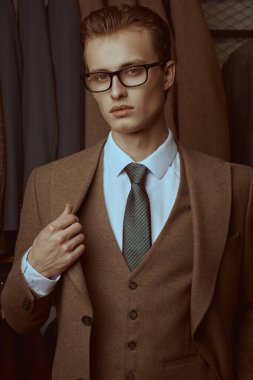 Zarif kahverengi takım elbiseli yakışıklı sarışın bir adam ve lüks klasik takım elbiseli bir mağazanın arka planında poz veren gözlüklü. Erkeklerin iş modası.