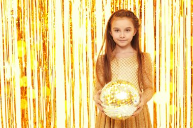 Altın bir gece elbisesi içindeki güzel küçük kız bir disko topuyla poz veriyor. Çocuk modası ve tatiller. Çocuk partisi. Fotokopi alanı ile şenlikli altın folyo perde arkaplanı. 