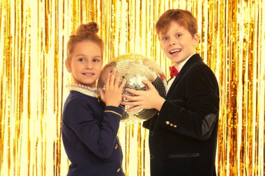 Çocuk partisi. Gece kıyafetleri içinde neşeli küçük bir kız ve oğlan, arka plandaki parlak altın folyo perdelerde bir disko topuyla neşeli bir partide poz veriyorlar. Çocuk modası ve tatiller. 