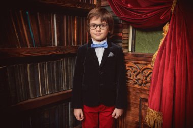 Zarif klasik okul üniforması ve gözlüklü yakışıklı modern çocuk lüks bir kütüphane içinde poz veriyor. Çocukların okul modası. Gözlük stili. 