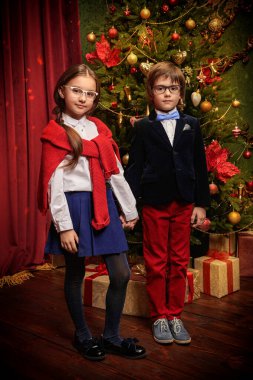 İki zarif, klasik okul üniformalı ve gözlüklü modern çocuk bir Noel ağacının yanında lüks bir iç mekanda poz veriyorlar. Noel süsü. Çocukların okul modası..