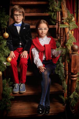 Çocukların okul modası. İki zarif, klasik okul üniformalı ve gözlüklü modern çocuk Noel süslemeleri olan lüks bir iç mekanda merdivenlerde oturuyorlar. Noel ve Yeni Yıl kavramı.