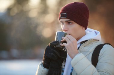 Örgü şapkalı ve ceketli yakışıklı genç adam ısınmak için sıcak bir içecekle kış parkında duruyor. Kışın açık hava aktiviteleri.