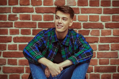 Kotlu ve flanel gömlekli yakışıklı genç bir adam tuğladan bir duvara yaslanmış oturuyor ve gülüyor. Gençlik modası. Mutlu insanlar ve duygular. Stüdyo resmi.