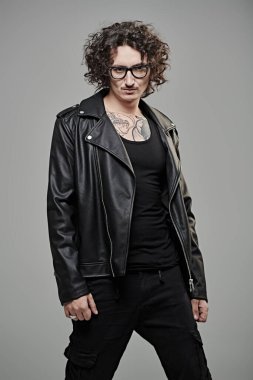 Siyah kıvırcık saçlı, vücudunda dövmeleri olan, siyah deri ceket ve gözlük takan gayri resmi bir rock müzisyeni. Gri arkaplanda stüdyo portresi.