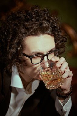 Siyah kıvırcık saçlı, zarif siyah takım elbiseli, beyaz gömlek ve gözlüklü heybetli bir adam viski içiyor. Geçmişe dönük bir portre. Zengin yaşam tarzı. Erkek modası.