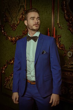 Erkekler için akşam modası. Beyaz gömlekli ve papyonlu zarif mavi takım elbiseli yakışıklı bir adam lüks bir dairede duruyor. İş biçimi.