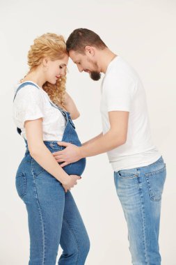 Sevgi dolu bir koca ve güzel hamile bir eş bebek bekliyor. Beyaz tişörtlü ve kot pantolonlu sevgili eşler göbeklerine sarılıyorlar beyaz bir stüdyo geçmişinde bir bebekle.