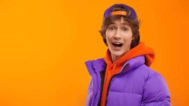 Parlak turuncu kapüşonlu, mor ceketli ve beysbol şapkalı mutlu bir gencin portresi. Fotokopi alanı olan sarı stüdyo. Gençlik Moda. 