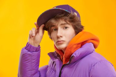 Parlak turuncu kapüşonlu, mor ceketli ve beysbol şapkalı modaya uygun bir genç. Sarı stüdyo geçmişi. Gençlik Moda. 