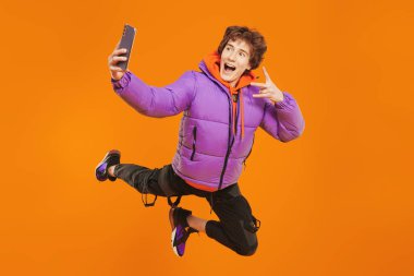 Duygusal ergen çocuk havada süpermen gibi uçuyor, bağırıyor ve telefonda selfie çekiyor. Turuncu arka plan. Modern gençlik kuşağı. Sosyal ağlar, internet toplulukları.